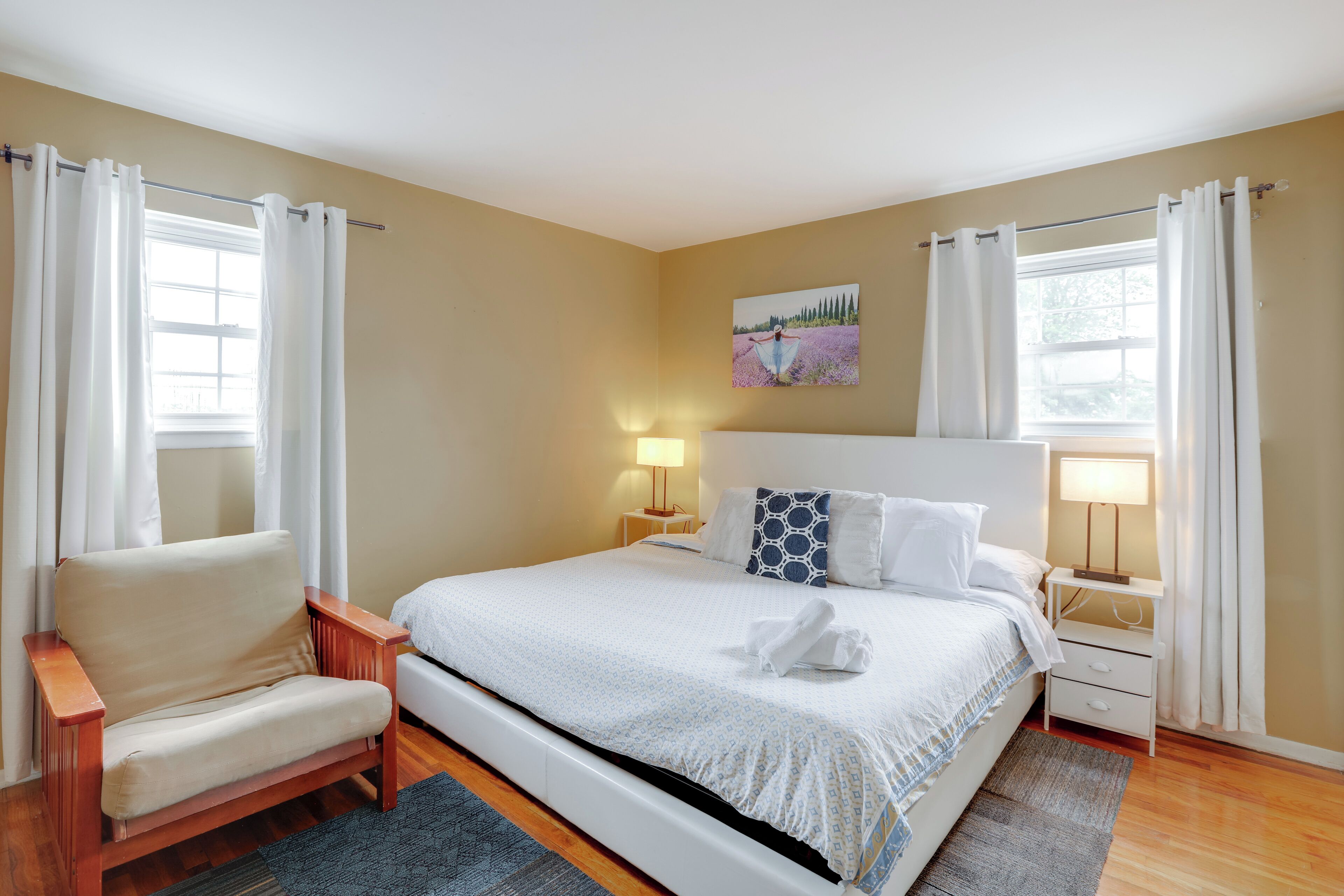 A neatly arranged bedroom with a bed, a chair, a nightstand, and a painting on the wall.