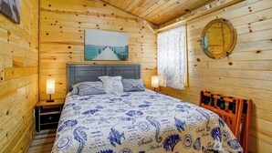 Une chambre dans une cabane en bois, avec un lit, des tables de chevet, un ventilateur de plafond, une fenêtre avec des rideaux et un miroir fixé au mur.