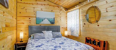 A wooden cabin bedroom with a bed, nightstands, a ceiling fan, a window with curtains, and a wall-mounted mirror.