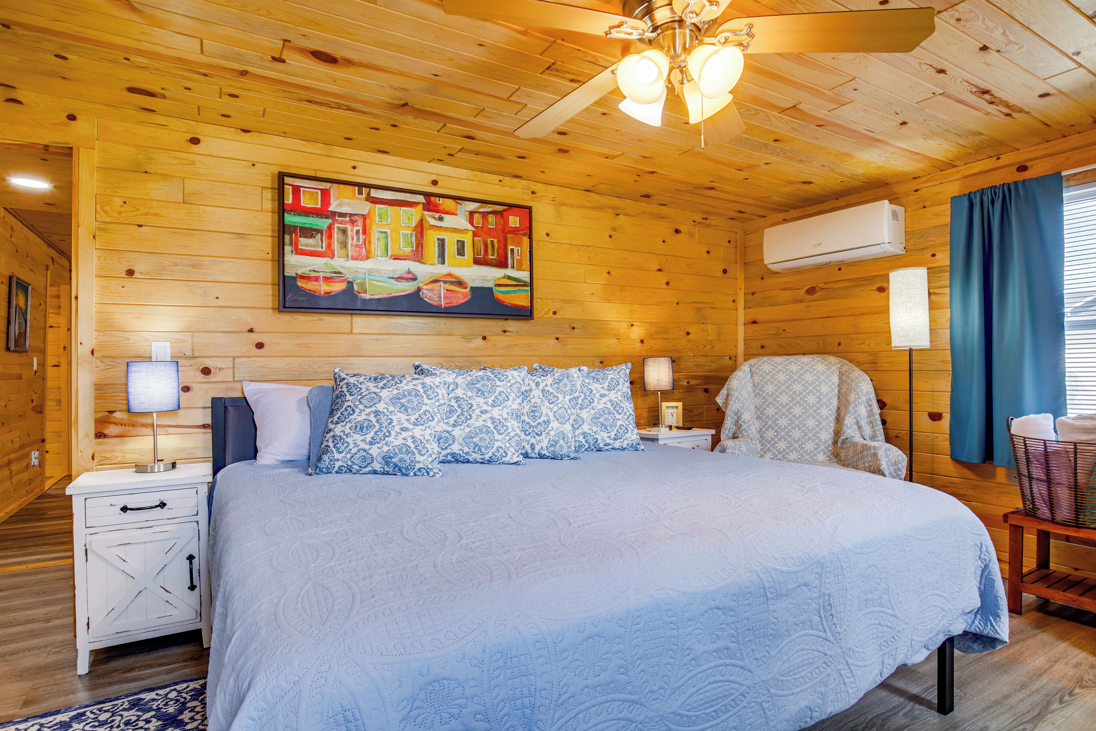 Une chambre de style cabane en bois, avec un grand lit, deux tables de chevet, un ventilateur de plafond et un tableau coloré accroché au mur.