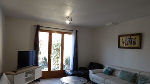 Ein Wohnzimmer mit Flachbildfernseher auf einem weißen Schrank, einem weißen Sofa mit grünen Kissen, einem kleinen runden Tisch und einer Schiebetür zu einem Außenbereich.