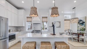 A modern kitchen with white cabinetry, stainless steel appliances, a central island with bar stools, and decorative wicker pendant lights. - Bird Feather 160tai - C (Port Aransas)