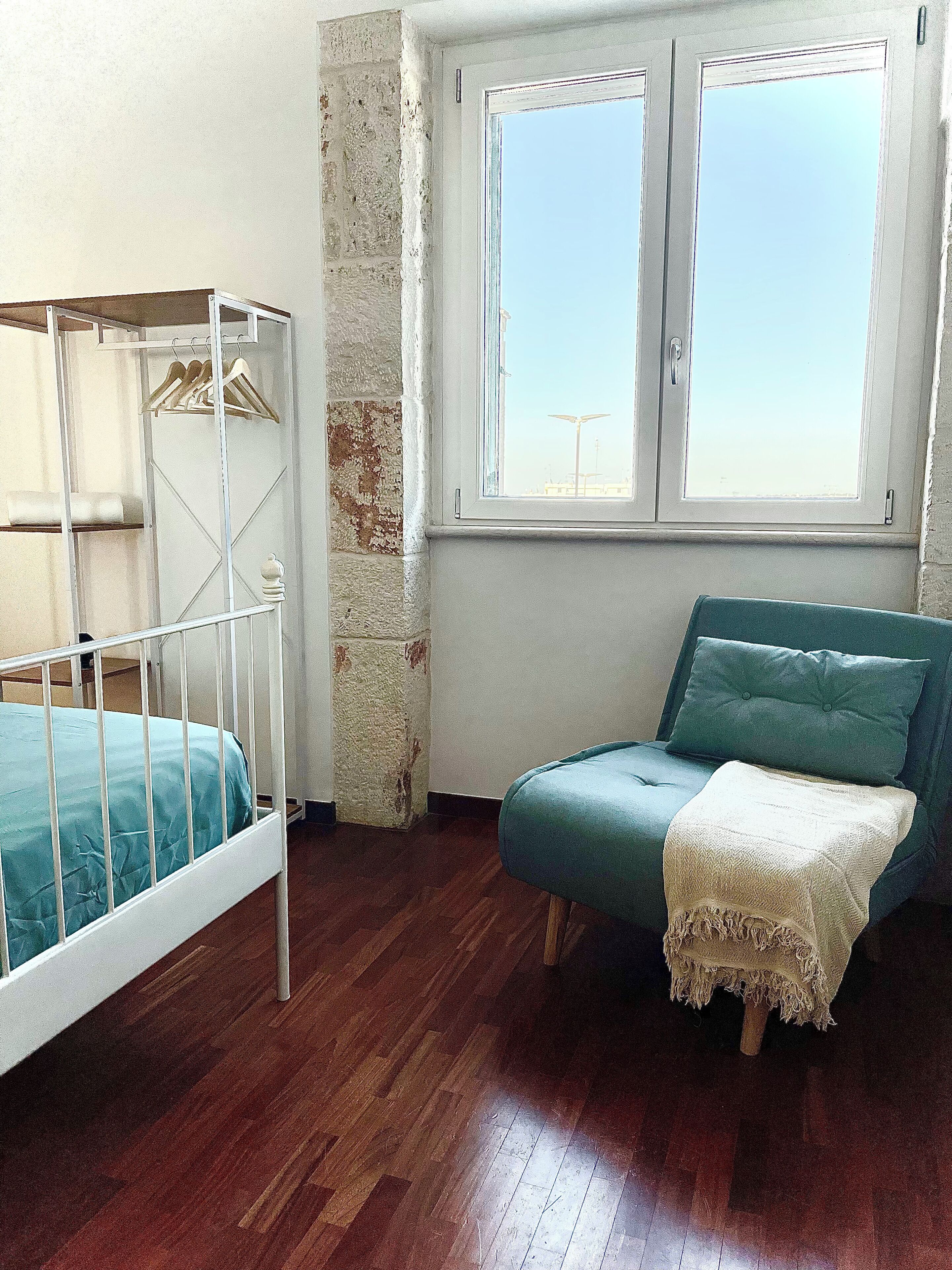 A room with a wooden floor, a white crib with a blue mattress, a teal sofa with a white throw blanket, and a window with a view of the sky.