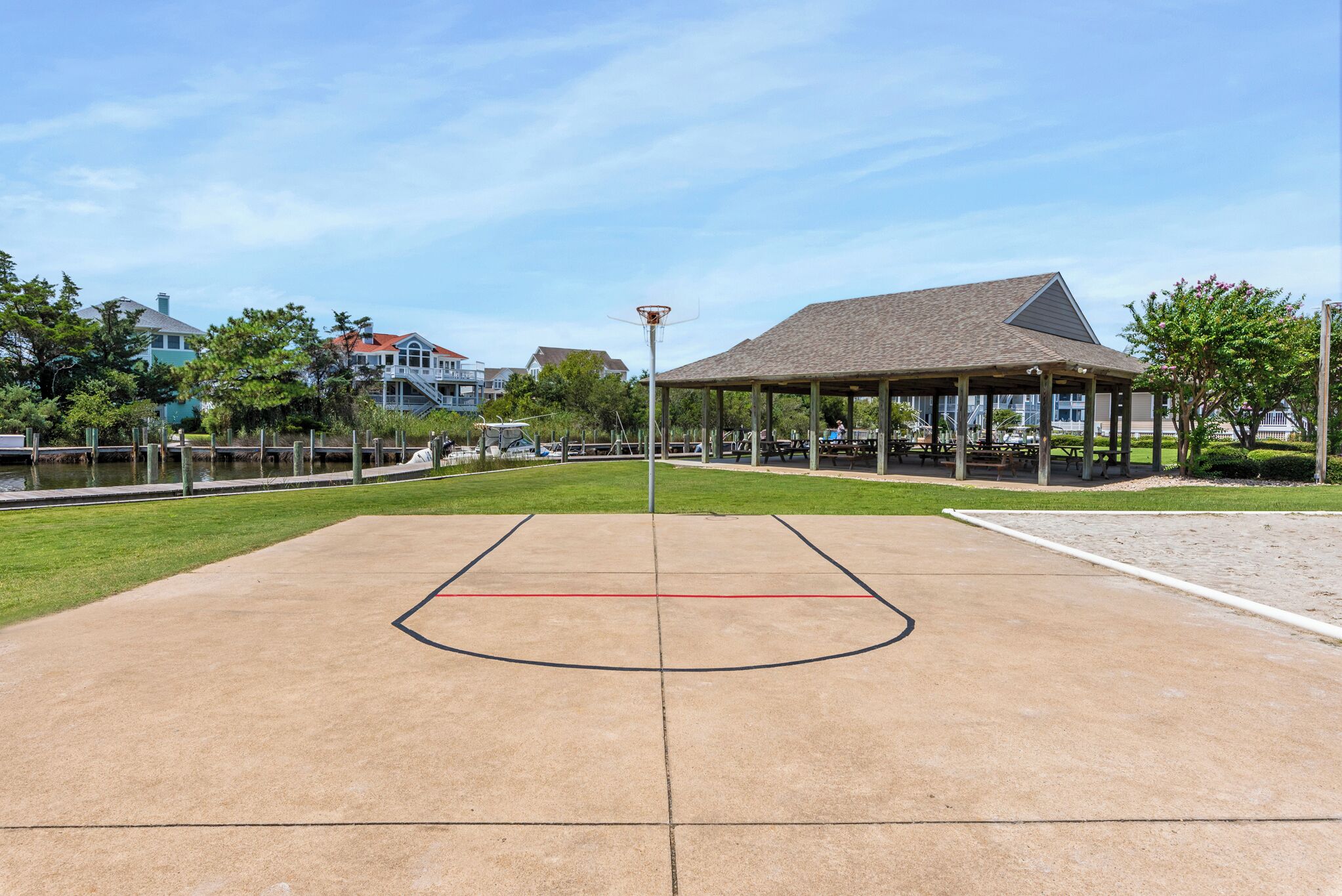 Una cancha de baloncesto al aire libre con una zona de asientos techada, rodeada de árboles y casas.