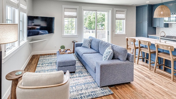 A modern living room with a gray sofa, wooden floor, and a kitchen area with blue cabinets and a hanging light fixture. - Carlton Landing-Carriage House (Carlton Landing)