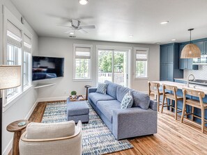 A modern living room with a gray sofa, wooden floor, and a kitchen area with blue cabinets and a hanging light fixture.