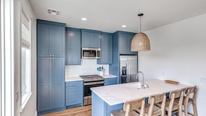A modern kitchen with blue cabinets, a stainless steel refrigerator, a microwave, and a large island with wooden chairs. - Carlton Landing-Carriage House (Carlton Landing)