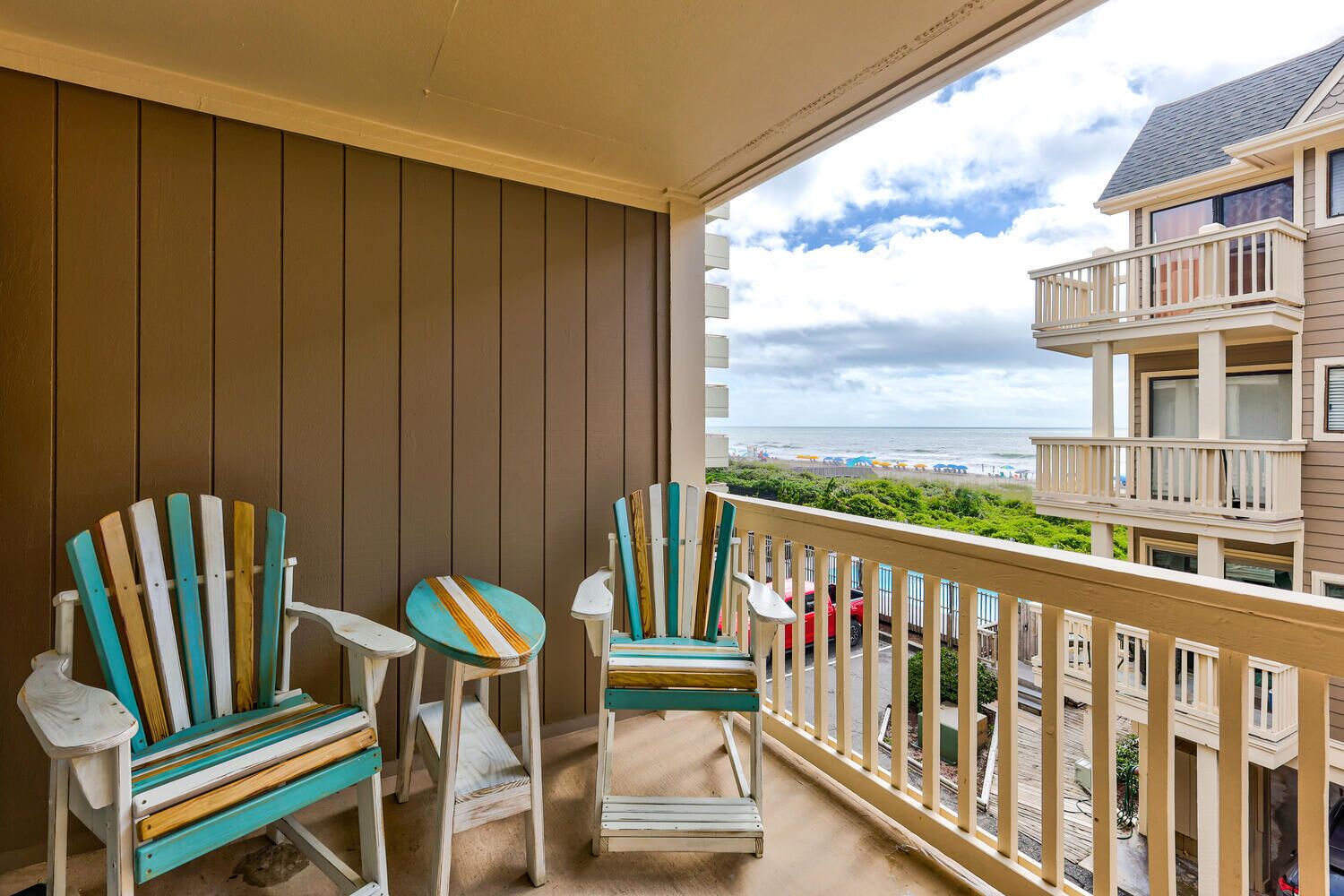 A balcony with two Adirondack chairs and a small table, overlooking a beach and a multi-story building.
