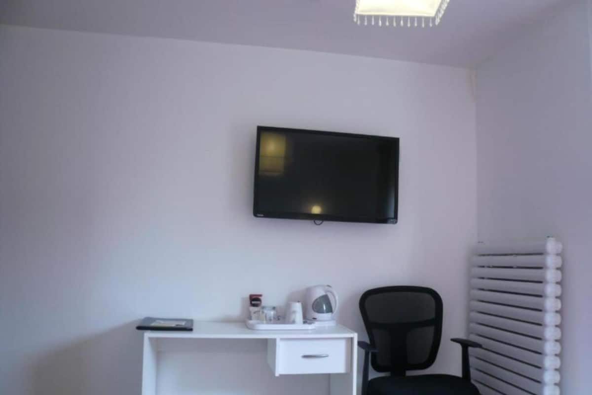 A small, clean room with a white desk, a black office chair, a flat-screen TV mounted on the wall, and a radiator on the right.