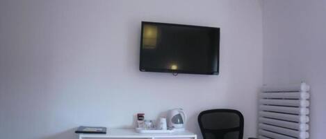 A small, clean room with a white desk, a black office chair, a flat-screen TV mounted on the wall, and a radiator on the right.