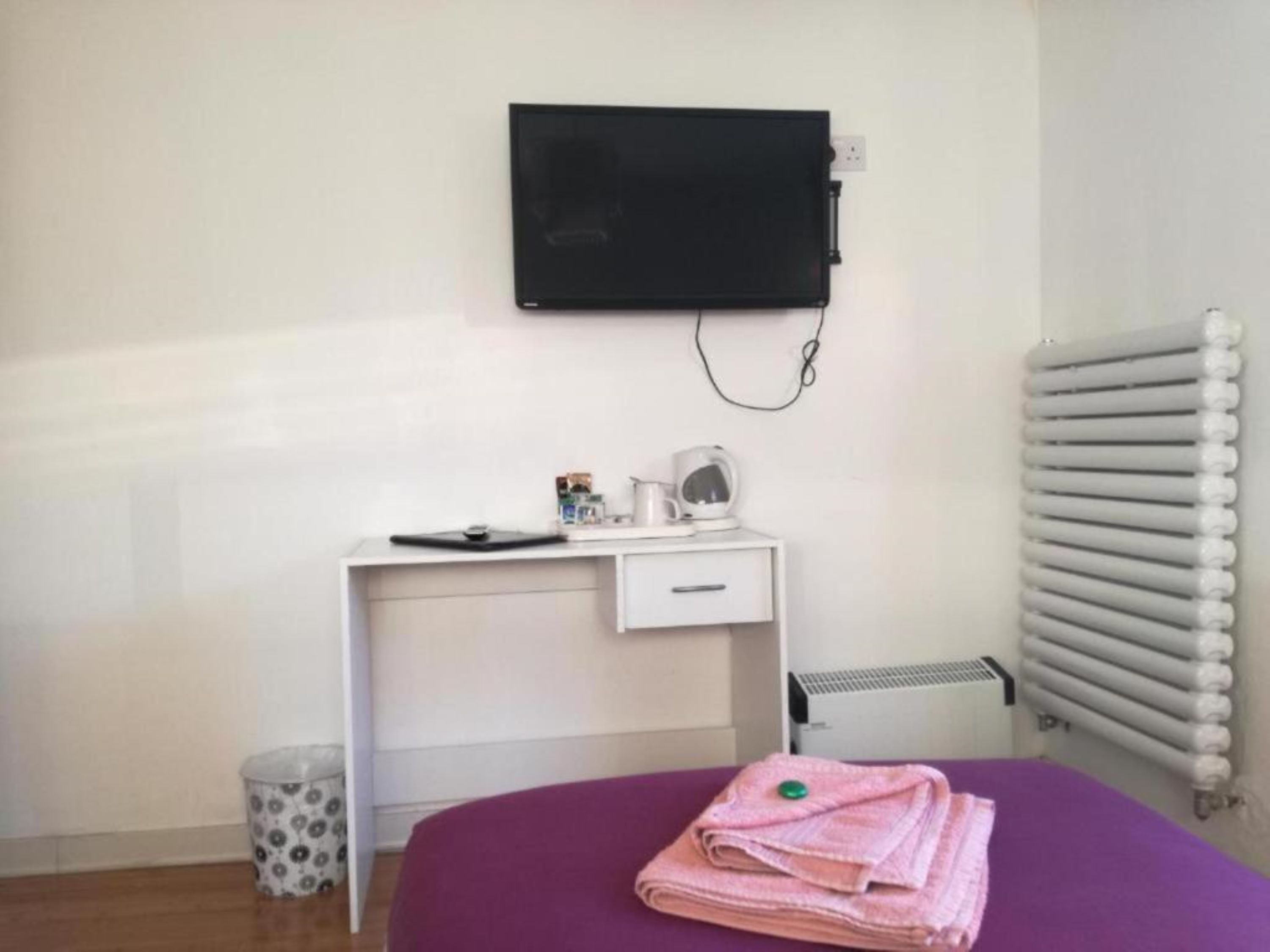 Une chambre comprenant un lit, un bureau avec un ordinateur, un radiateur et un téléviseur fixé au mur.