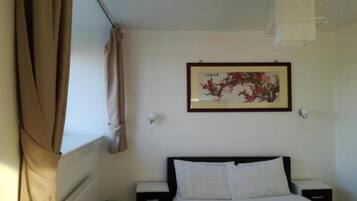 A hotel room with a bed, two bedside tables, a radiator, a window with curtains, and a framed picture on the wall.