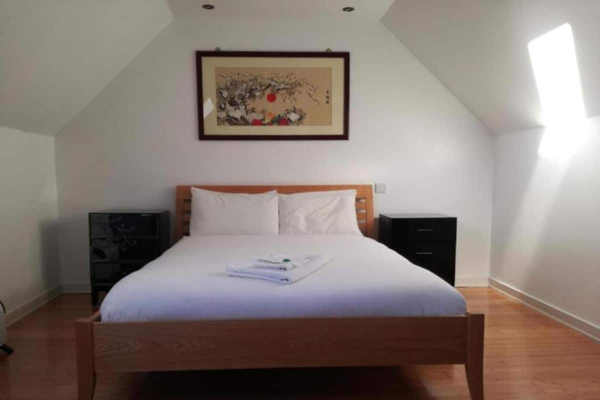 A bedroom with a wooden bed, white bedding, a framed artwork on the wall, and a nightstand with a lamp.