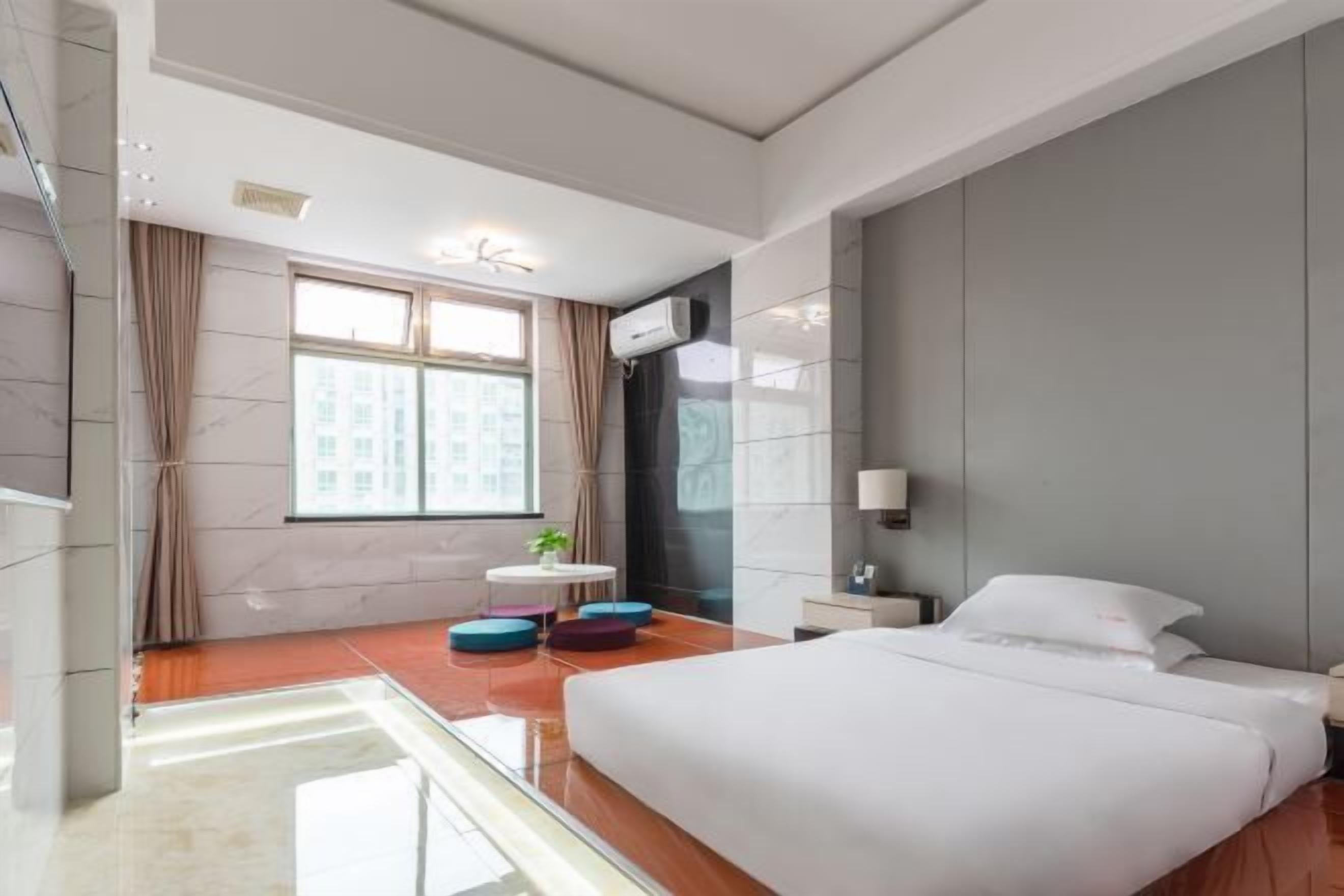 A modern hotel room with a large bed, a small round table with colorful stools, a window with curtains, and a view of buildings outside.
