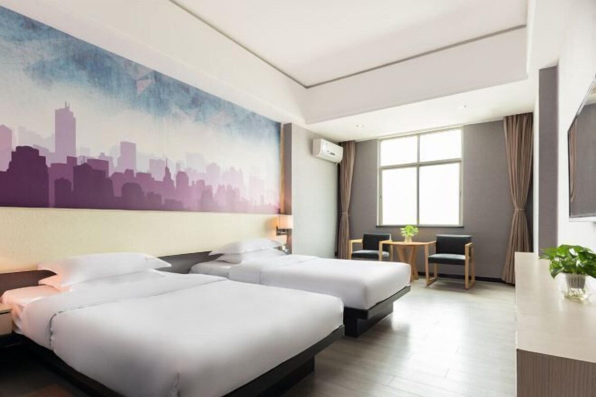 A hotel room with two beds, a cityscape mural, a large window, a desk, and a chair.