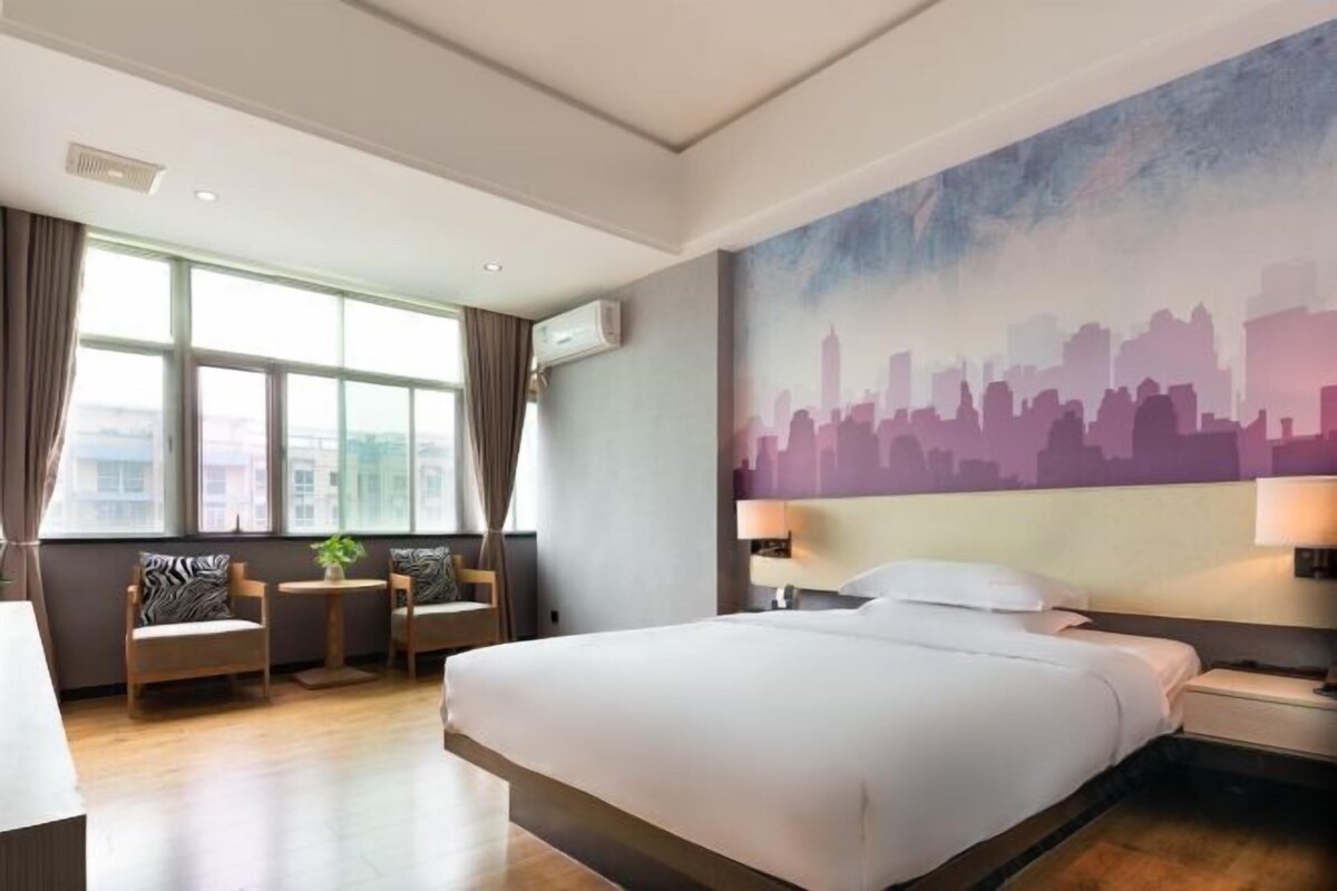 A hotel room with a large bed, a cityscape mural, a seating area with chairs and a table, and large windows.