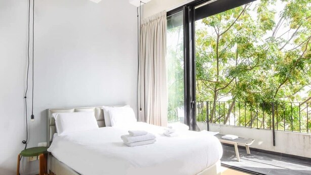 A modern hotel room with a large bed, a small bedside table, a window with a view of trees, and a power outlet on the wall. - Sky 2BR Penthouse by HolyGuest (Tel Aviv)