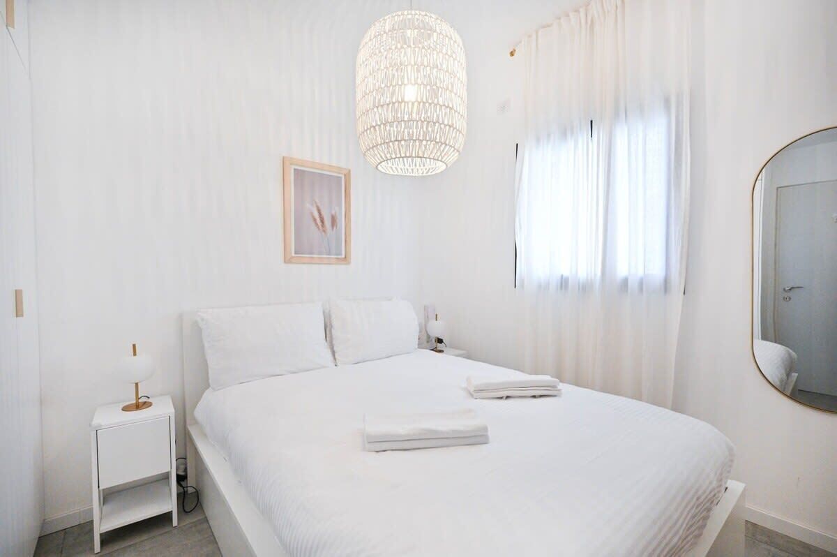 A clean, white hotel room with a large bed, a bedside table with a lamp, a hanging light fixture, and a mirror on the wall.