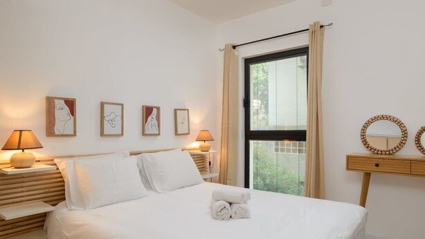 Een hotelkamer met een bed, bedlampen, kunst aan de muur, een raam met gordijnen, en een klein houten nachtkastje met een spiegel.