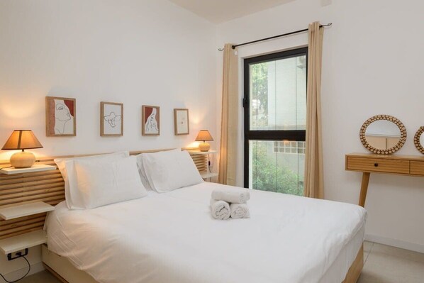 A hotel room with a bed, bedside lamps, artwork on the wall, a window with curtains, and a small wooden side table with a mirror.