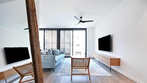 A modern living room with a sofa, two wicker chairs, a wooden bench, and a flat-screen TV mounted on the wall.
