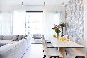 En moderne stue med sofa, spisebord og stole. Et vindue med lette gardiner og udsigt til bygninger.