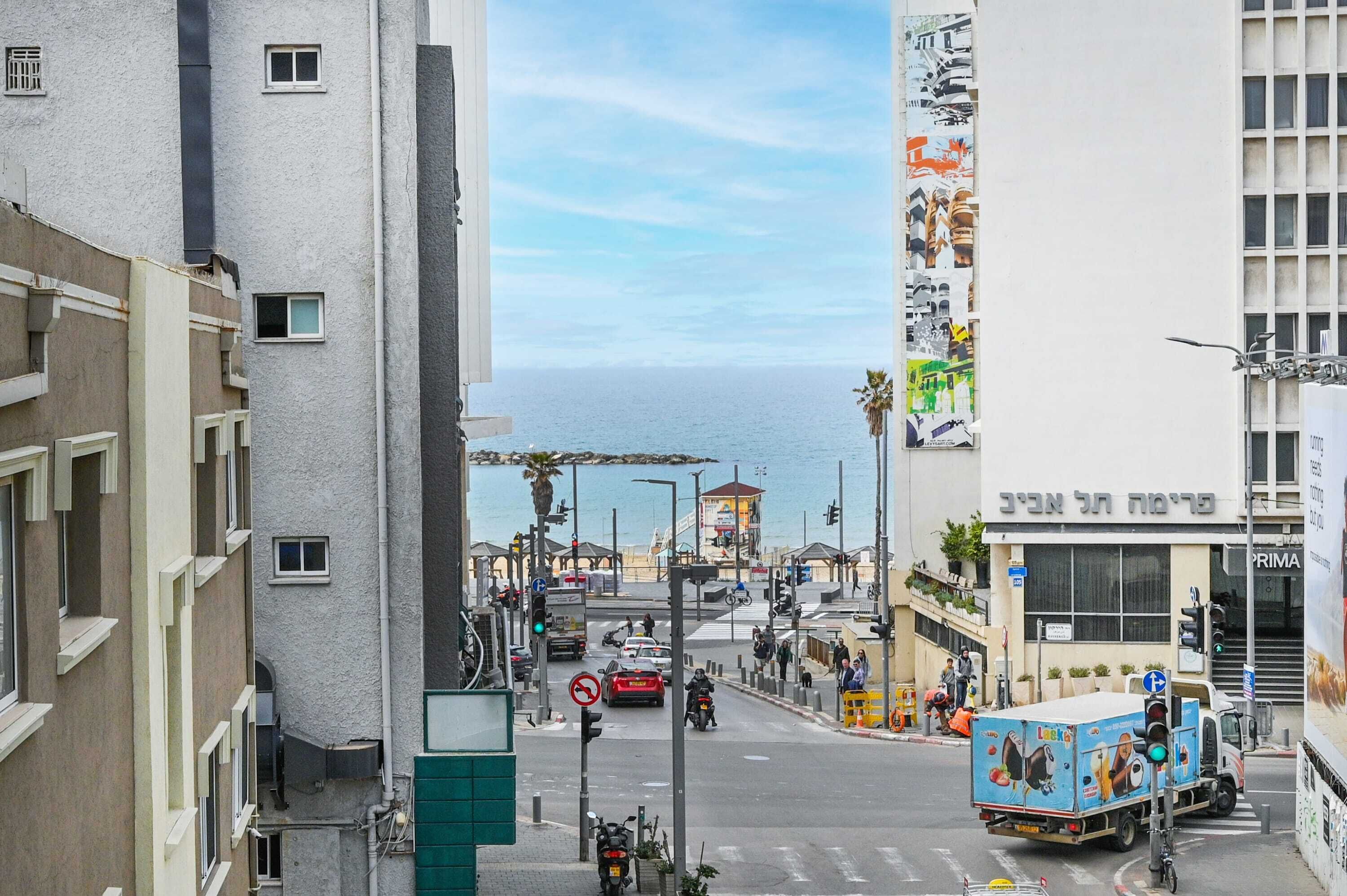 A street view with a building featuring a large poster on its side, a parked delivery truck, and a clear view of the sea in the background.