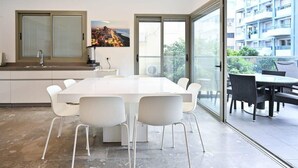 A modern kitchen with a large island, white chairs, and a dining area with a glass door leading to a balcony. - Mamma Mia 3BR in Frishman beach by HolyGuest (Tel Aviv-Yafo)