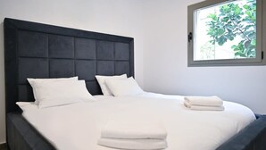 A neatly made bed with white linens and pillows, a dark headboard, and a window with a view of greenery. - Mamma Mia 3BR in Frishman beach by HolyGuest (Tel Aviv-Yafo)