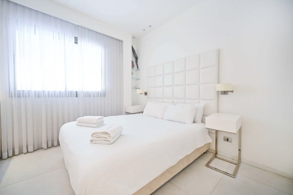 A modern hotel room with a large bed, white linens, a headboard with a light panel, bedside lamps, and a view of the city through sheer curtains.