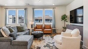A modern living room with a sectional sofa, a leather armchair, a white armchair, a black ottoman, a floor lamp, and a large window with a view of neighboring houses. - Desert Beach Retreat: Desert Color Lagoon + Beaches (St. George)