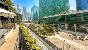 A modern urban plaza with a glass building, outdoor seating, and a landscaped area with trees. - HiGuests - Ahad Residences (Madinat Zayed)