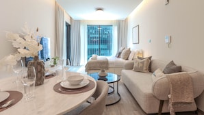A modern living room with a dining area, a sofa, and a view of the city through large windows. - HiGuests - Ahad Residences (Madinat Zayed)