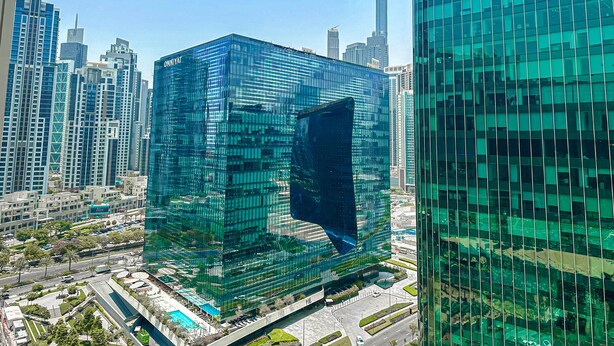 A modern glass building with a unique angular design, surrounded by other high-rise structures in a cityscape. - HiGuests - Ahad Residences (Madinat Zayed)