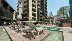 A modern hotel pool area with sun loungers, umbrellas, and a view of high-rise buildings. - HiGuests - Ahad Residences (Madinat Zayed)