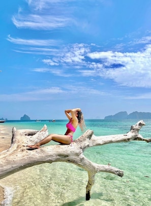 Una persona sentada sobre un tronco de madera a la orilla de un mar turquesa cristalino, con islas al fondo.