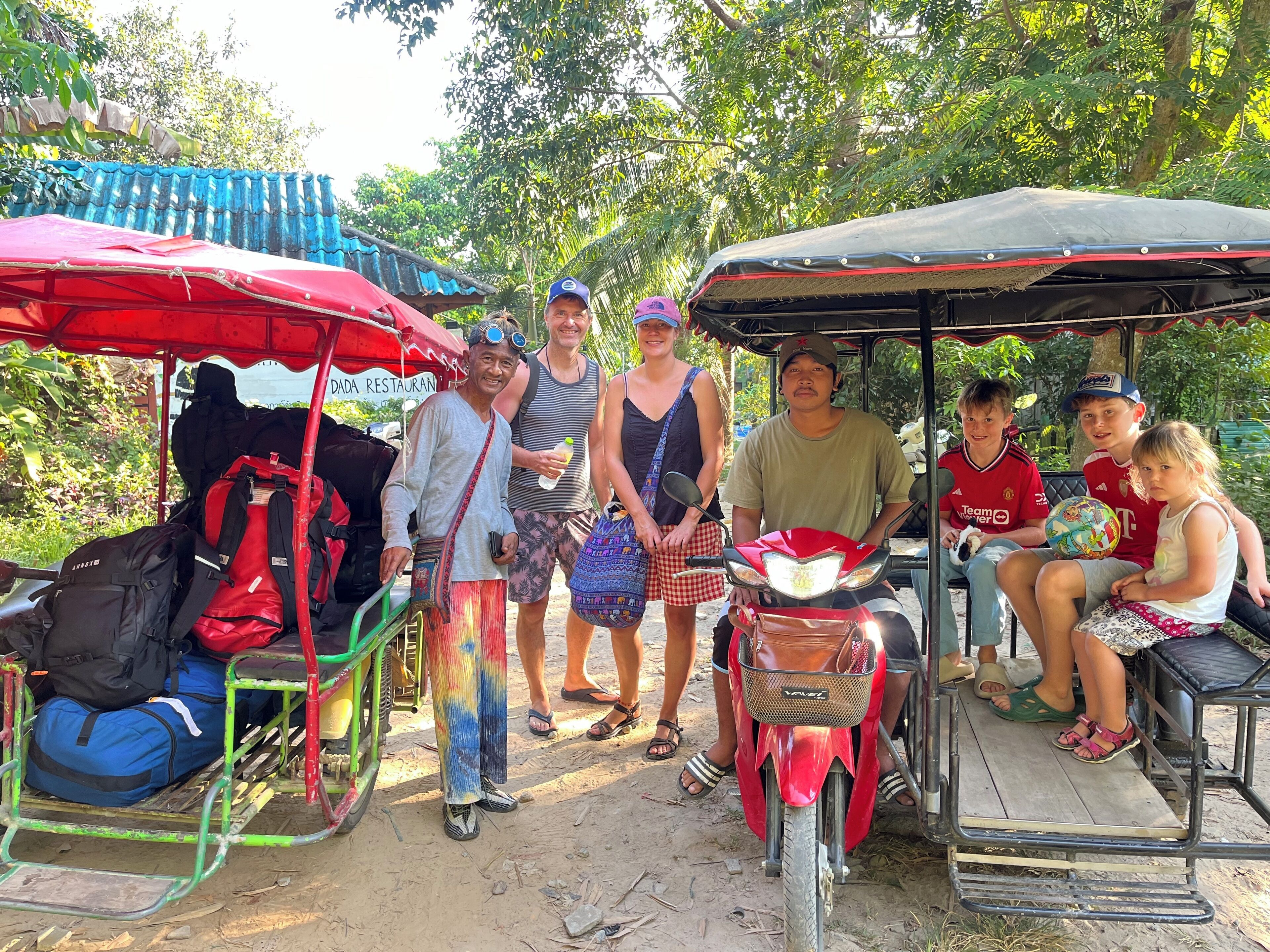 A group of people gathered around a red motorcycle and a cart with a red canopy, with trees and a building in the background.