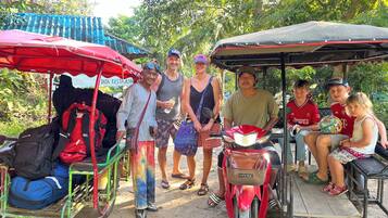 A group of people gathered around a red motorcycle and a cart with a red canopy, with trees and a building in the background.