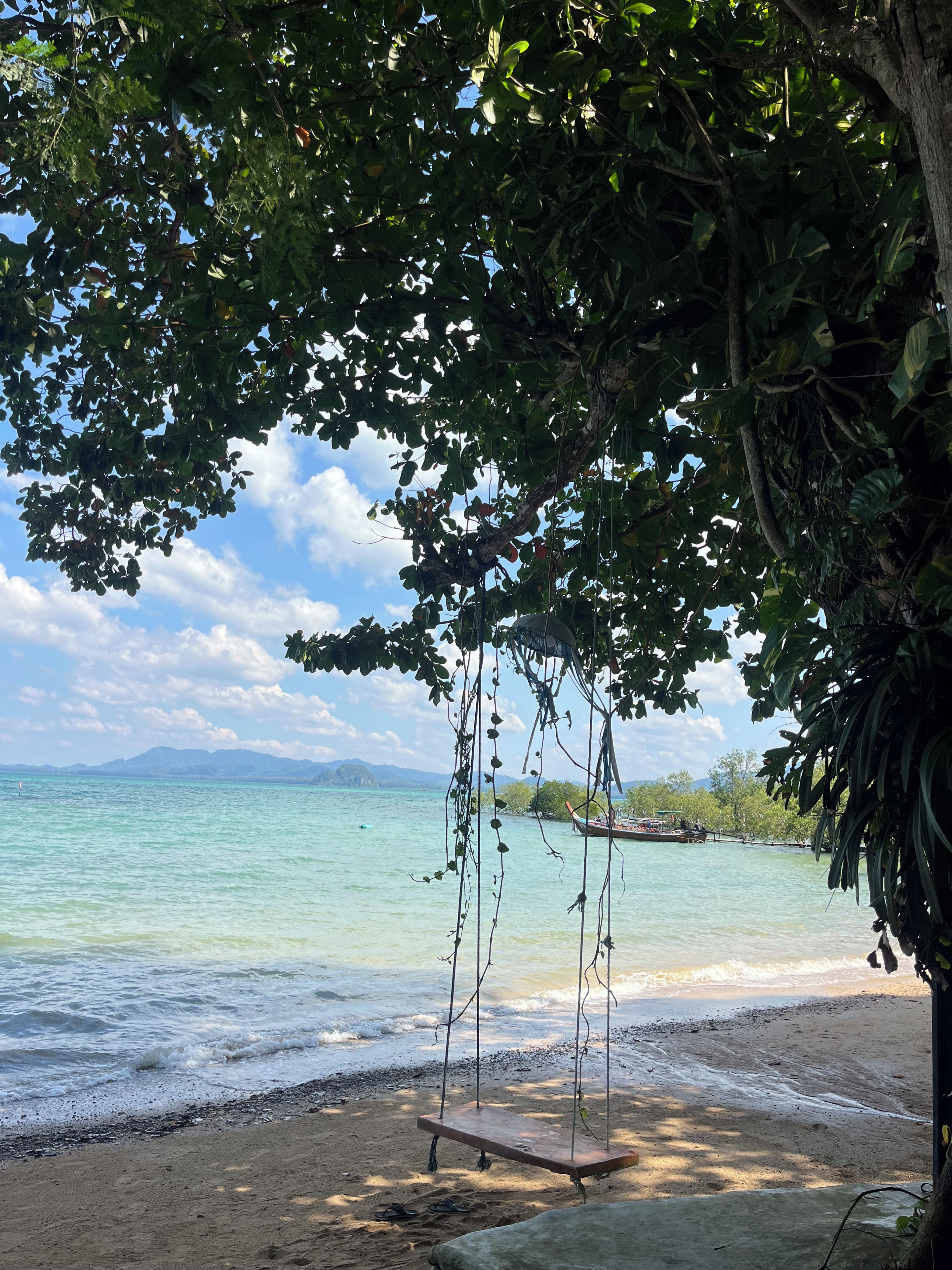 Una platja amb un bressol sota un arbre, aigua cristal·lina i muntanyes a la distància.