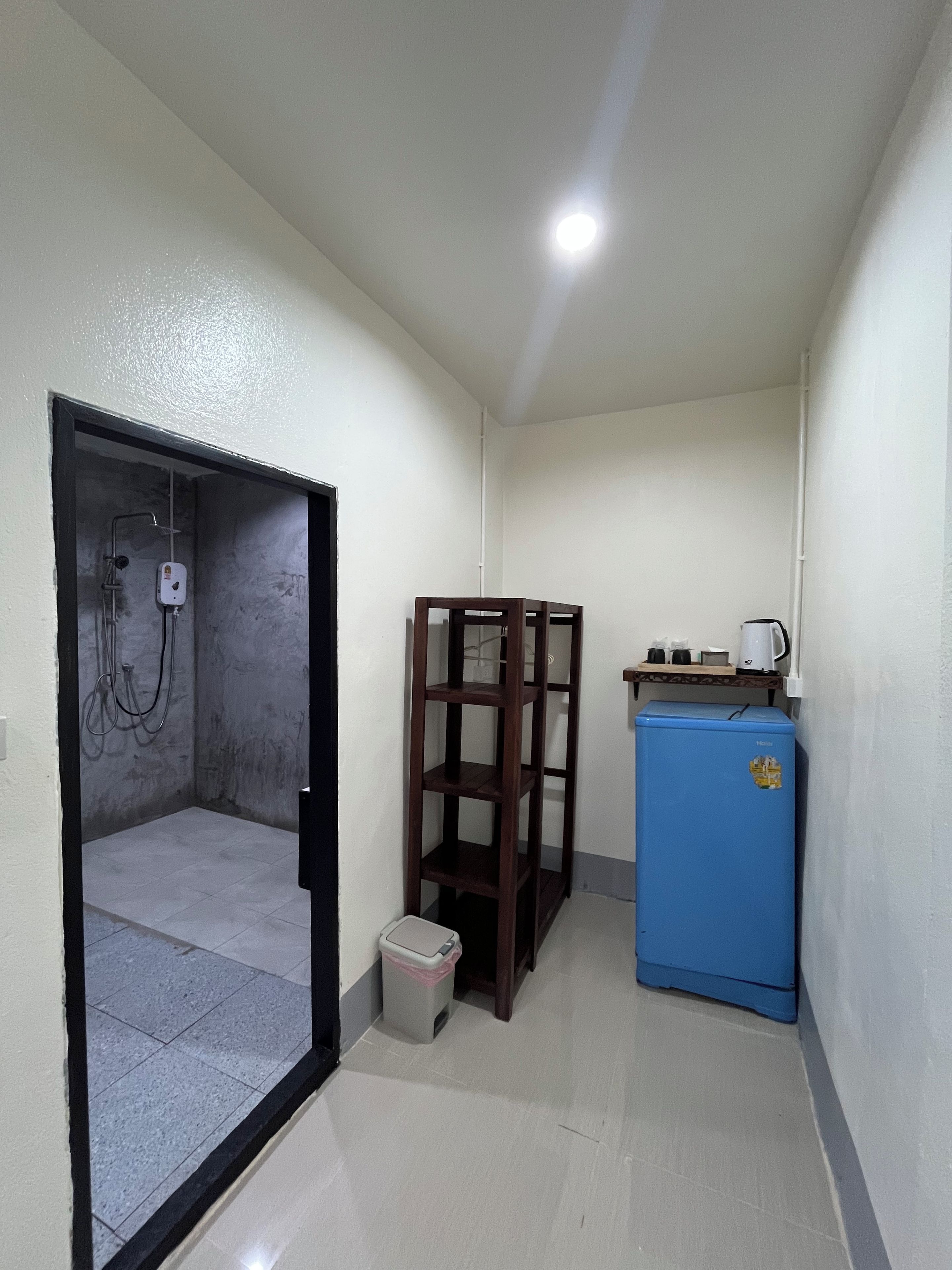 A narrow hallway with a shower area on the left, a wooden shelf unit, and a blue refrigerator.