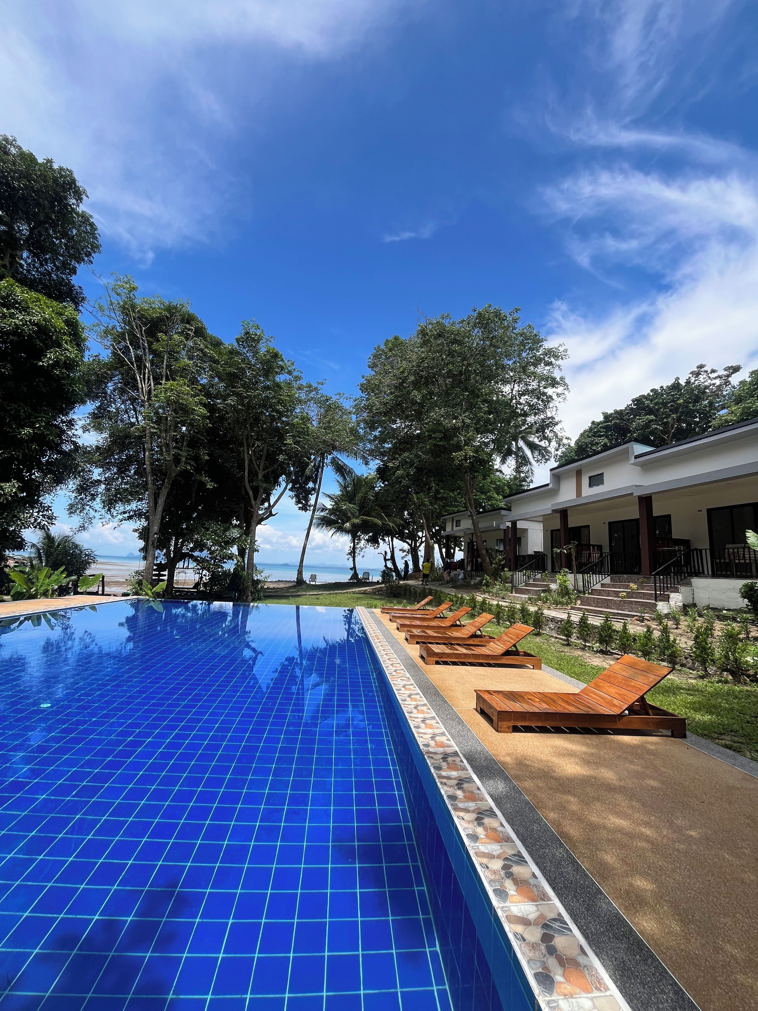 A swimming pool with a tiled edge, wooden lounge chairs, and a building with a balcony overlooking the pool.