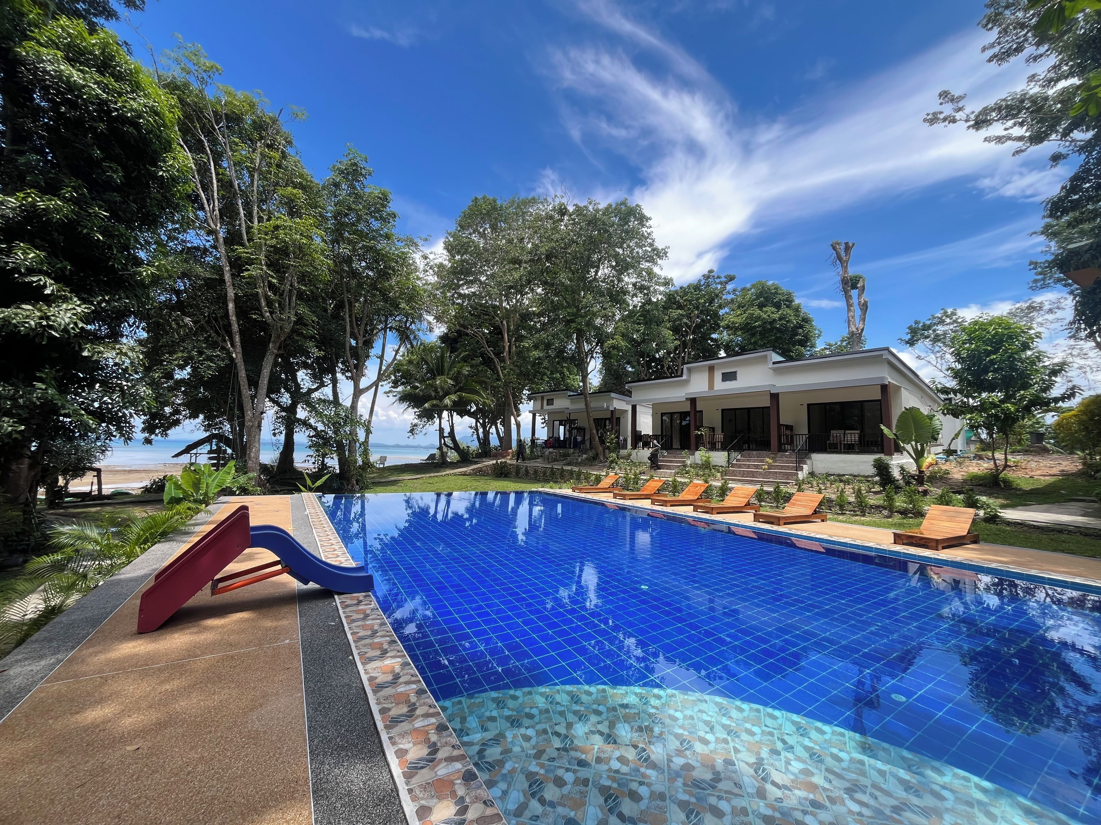 A swimming pool with a slide, surrounded by trees and a building with a covered patio.