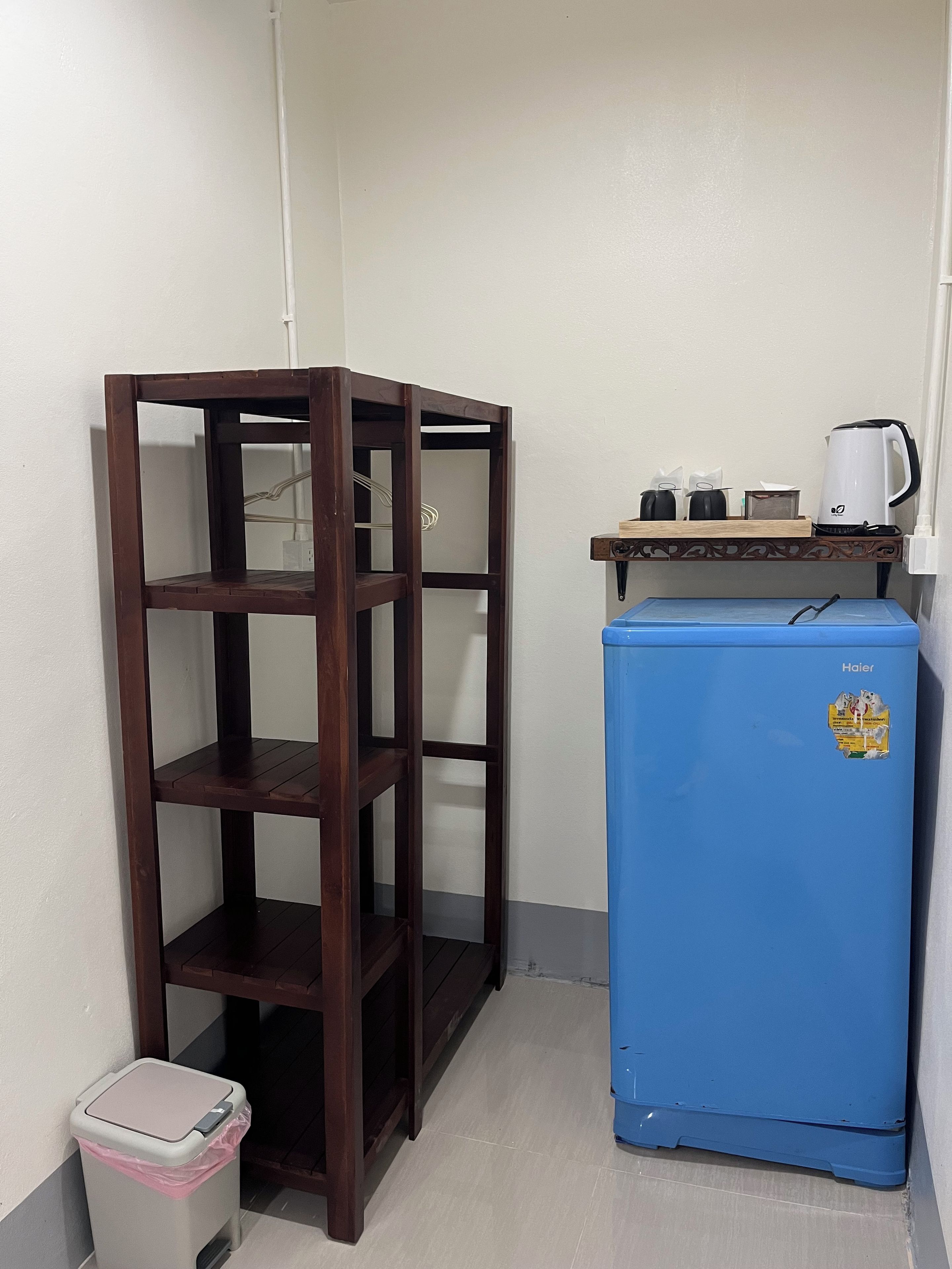 A compact room with a wooden shelving unit, a blue refrigerator, a small trash bin, and a shelf with a kettle and cups.