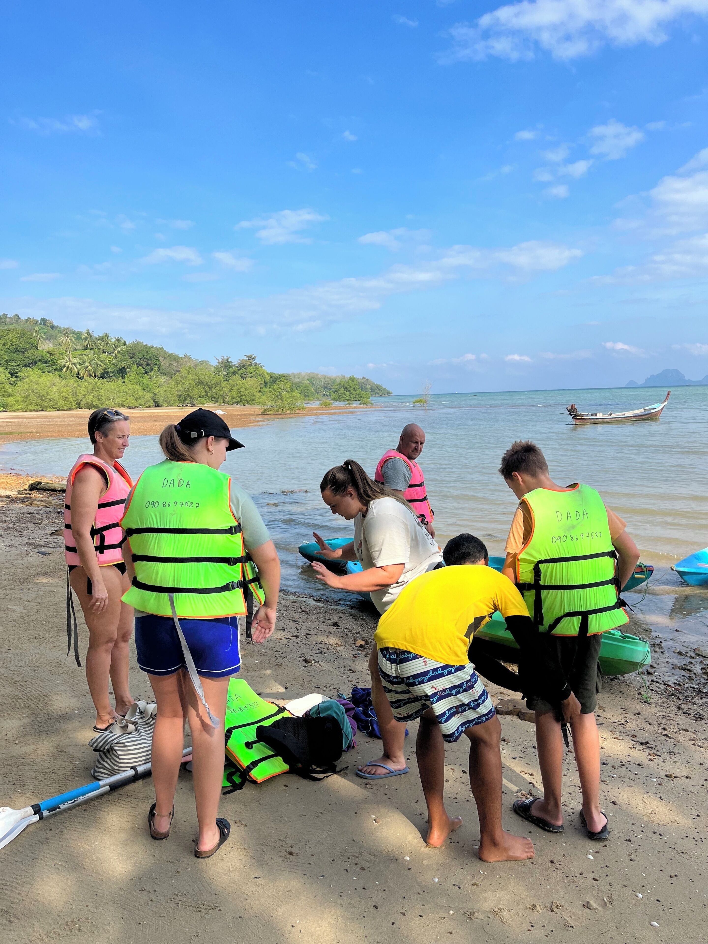 Un grupo de personas en una playa con chalecos salvavidas, un barco en el agua y un cielo despejado.