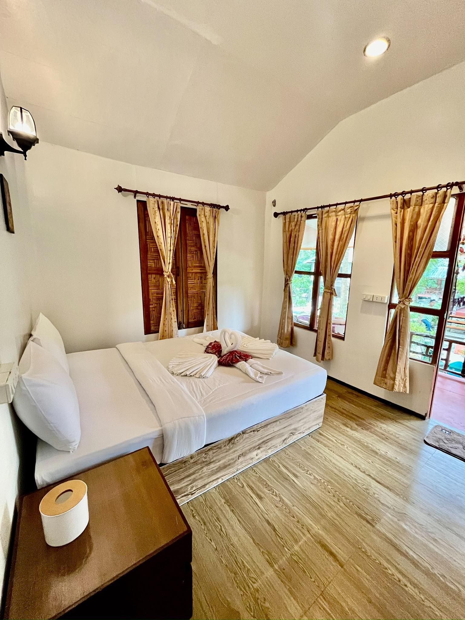 A hotel room with a bed, curtains, a wooden floor, and a nightstand with a tissue box.