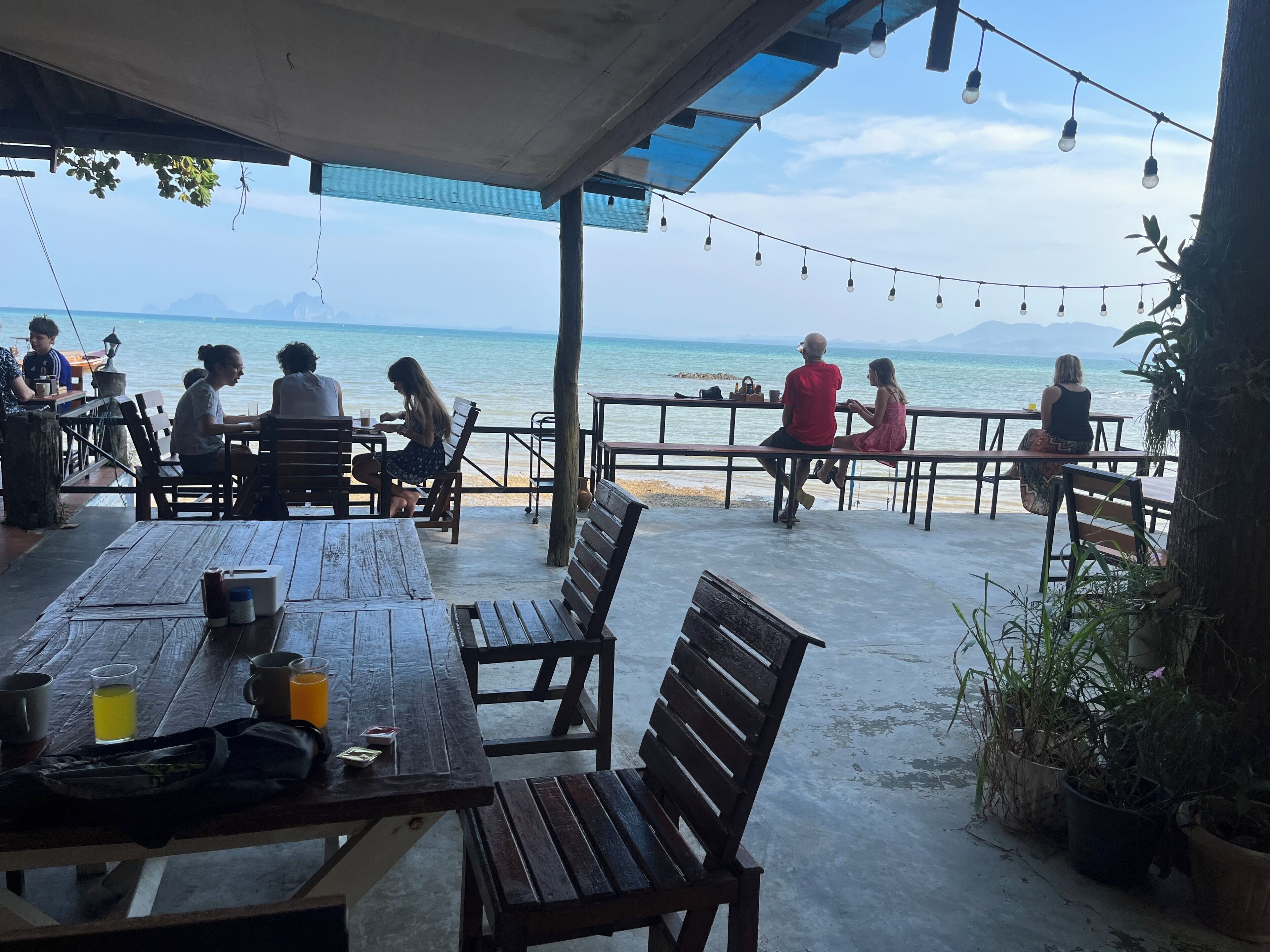 An outdoor seating area with wooden tables and benches, overlooking a beach and the sea.
