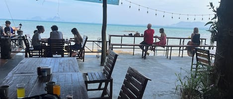 Un área de estar al aire libre con mesas y bancos de madera, con vista a una playa y al mar.