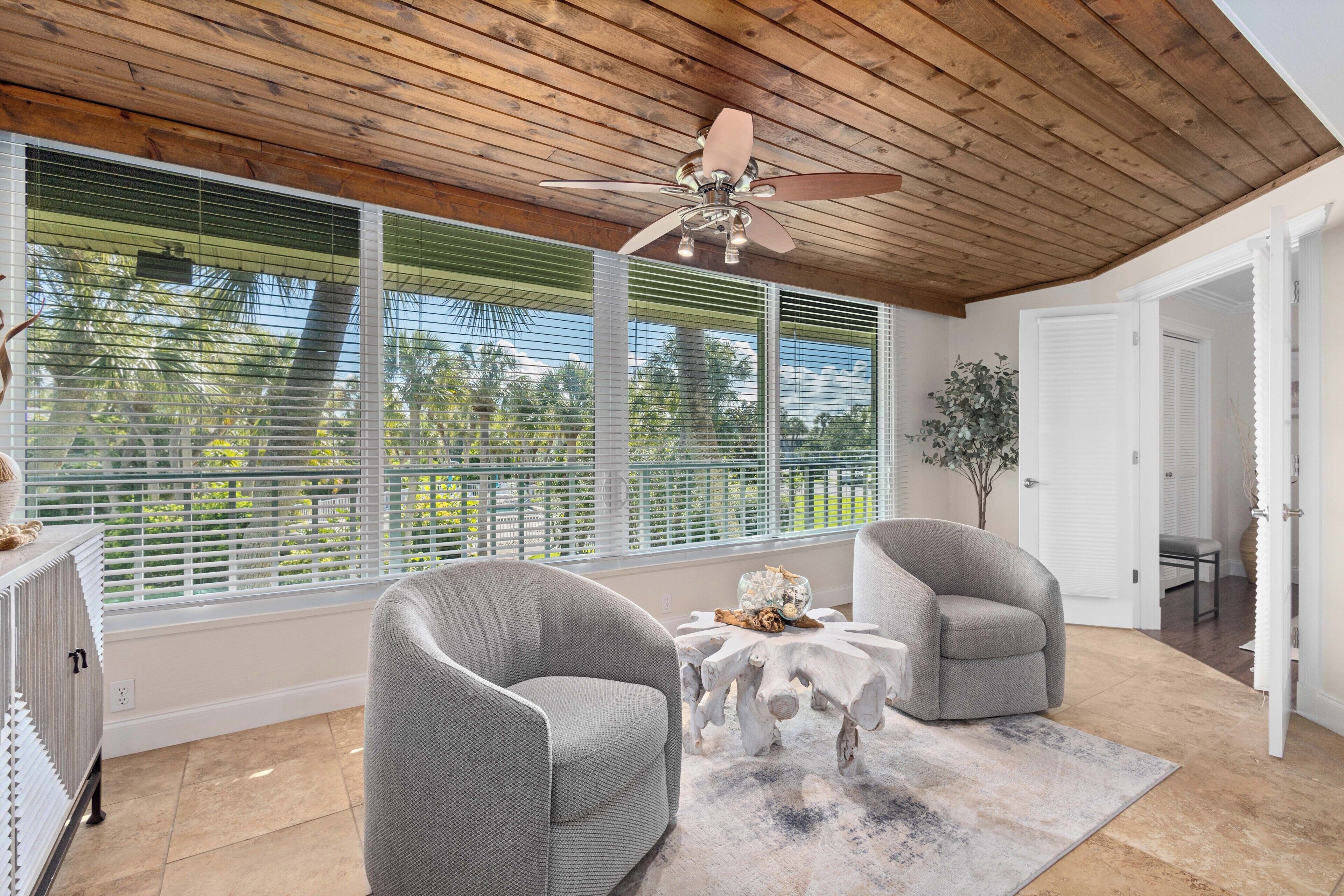 A room with a ceiling fan, two grey armchairs, a wooden coffee table, and large windows with blinds.