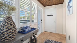 Une pièce avec un plafond en bois, une porte blanche, une grande fenêtre avec des stores, un vase décoratif et un plateau bleu contenant des boîtes.