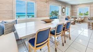 Een ruime eetruimte met een grote tafel en stoelen, grote ramen met uitzicht op zee, en een moderne kroonluchter.